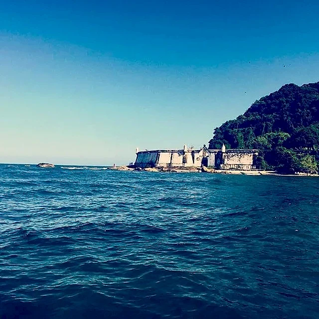 Mar agitado diante de uma pequena construção à beira da praia, cercada por mata densa em uma encosta. A cena é iluminada por luz diurna intensa, sob céu azul limpo.