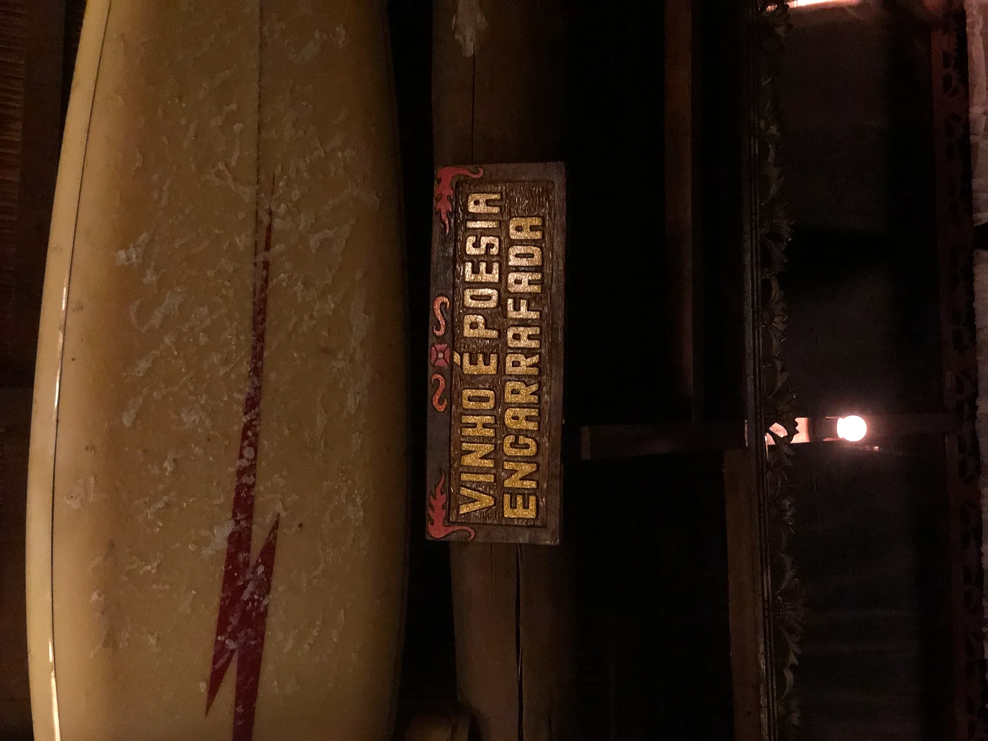 Prancha de surfe apoiada junto à entrada de uma pousada, com placa de boas-vindas em madeira. Ambiente interno rústico, iluminado por luz quente e baixa.
