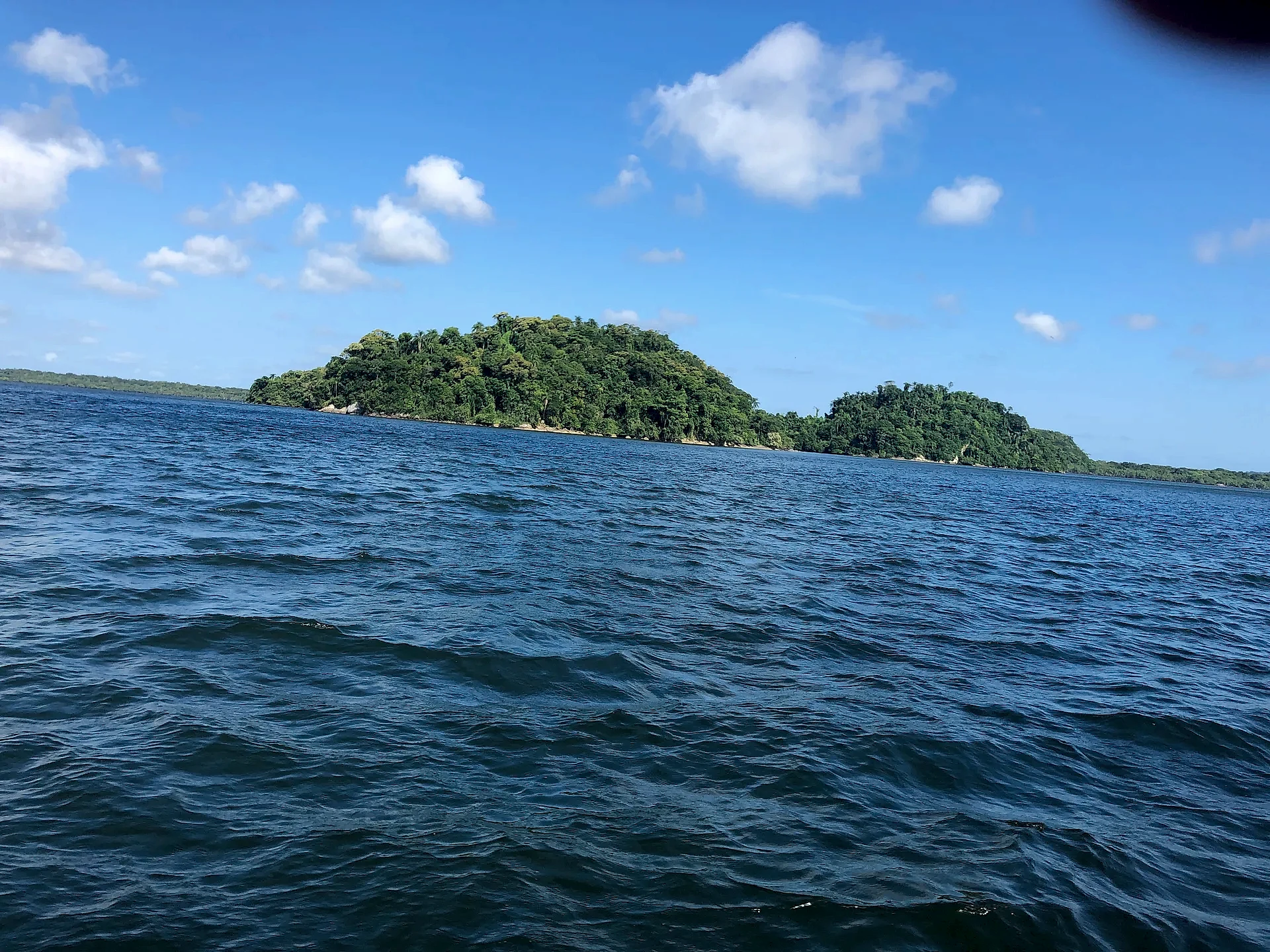 Vista do mar para uma pequena ilha coberta por mata densa, com ondulações em primeiro plano. O céu azul com nuvens esparsas e a luz diurna intensa destacam o verde da vegetação.