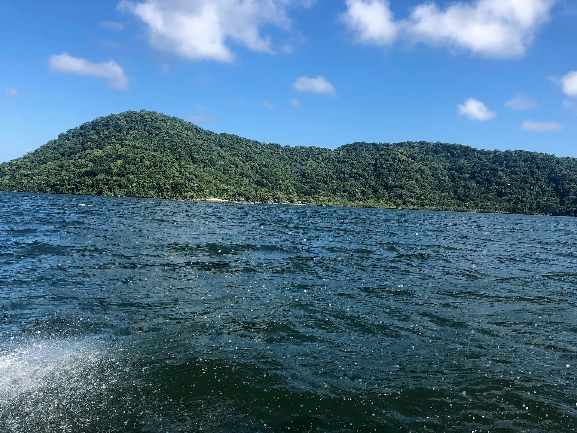 Mar com ondas em primeiro plano diante de morros cobertos por mata densa, vistos a partir de uma embarcação. O ambiente é aberto e natural, sob luz forte de dia ensolarado e céu azul com nuvens esparsas.