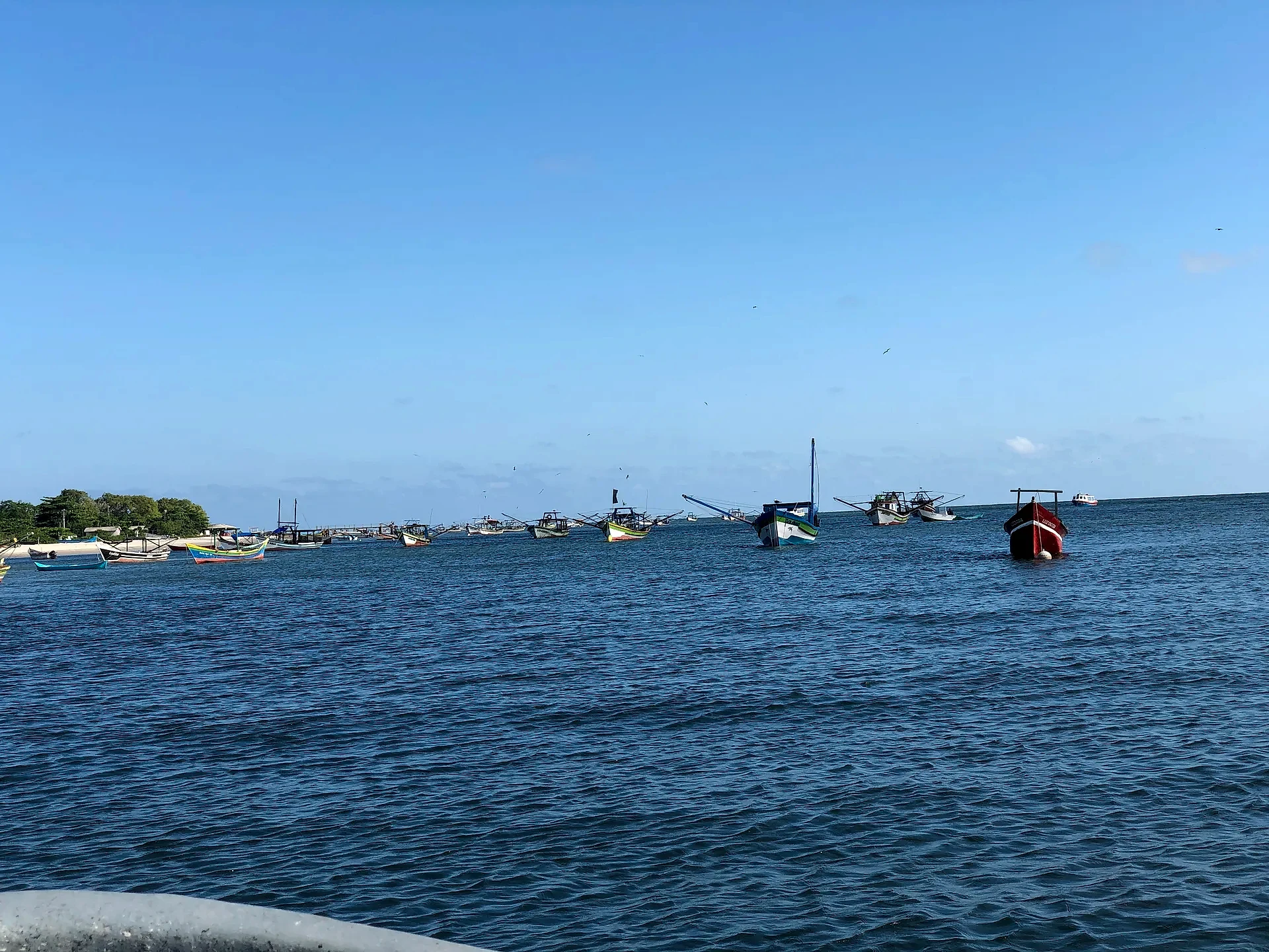 Barcos de pesca coloridos estão ancorados em águas calmas, próximos a uma faixa de mata na margem. O céu amplo e azul, sob luz clara do dia, reforça a atmosfera tranquila do litoral.