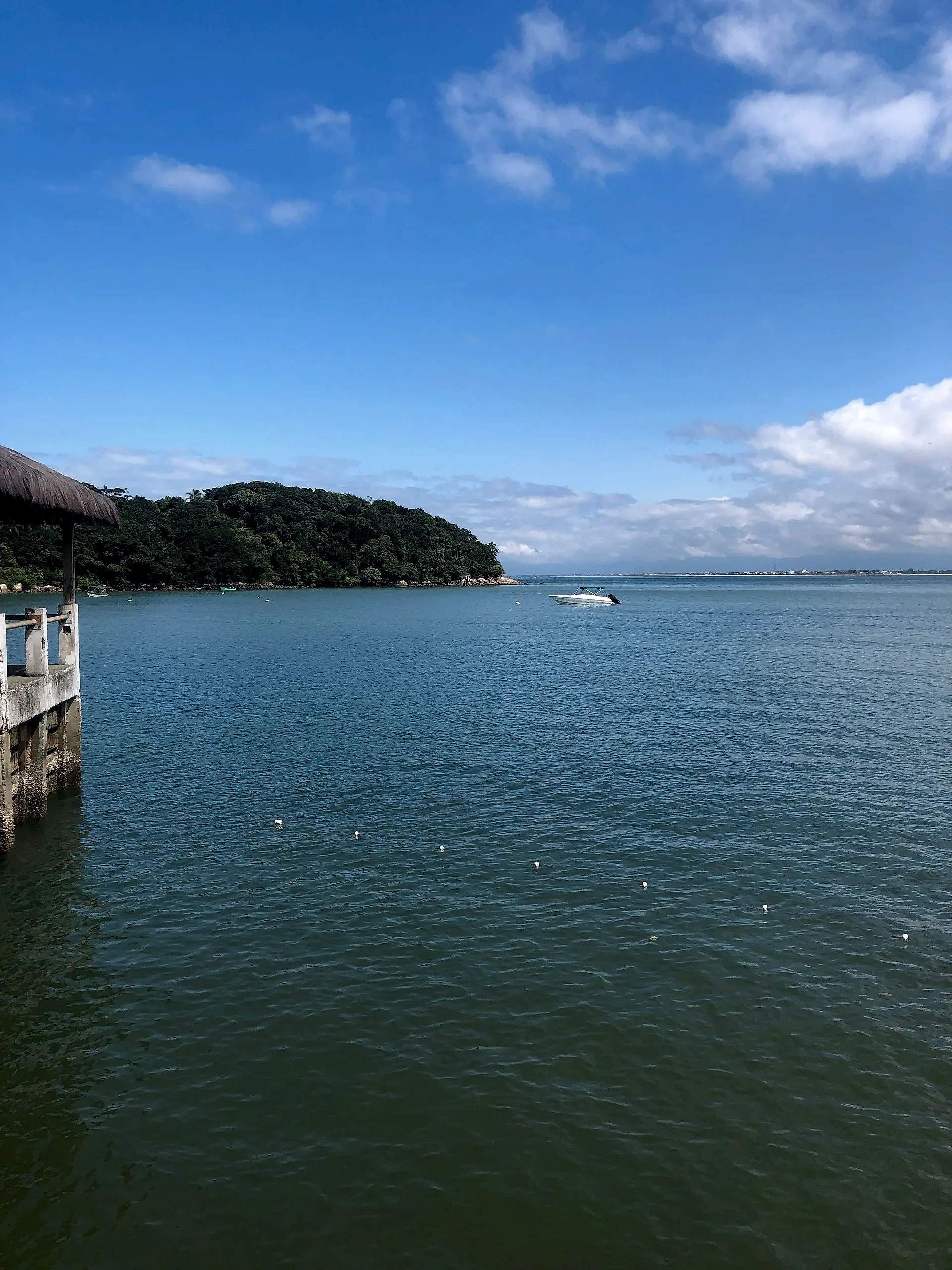 Vista de águas calmas junto a um trapiche, com barco ancorado e morro coberto pela mata ao fundo. A luz diurna intensa realça o azul do céu e os reflexos na superfície da baía.