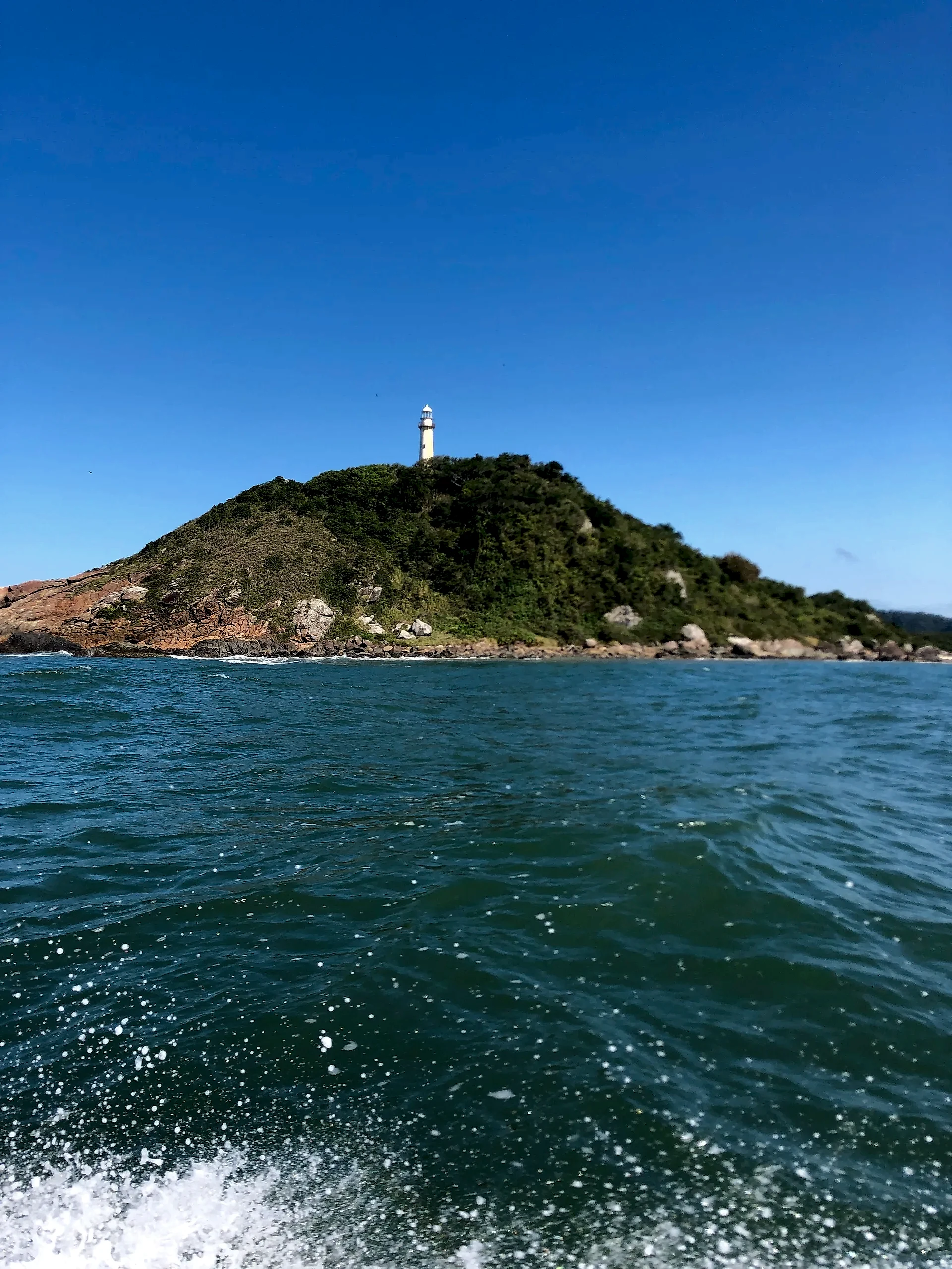 Farol sobre morro coberto pela mata, com costão rochoso à beira-mar visto a partir de um barco. A luz intensa do dia realça o céu azul, a água verde-azulada e a espuma das ondas.