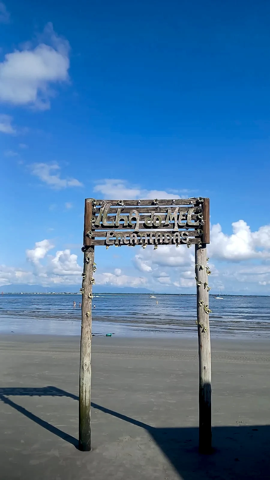 Placa rústica de madeira na areia indica Encantadas, diante de uma praia de águas calmas e pequenos barcos ao fundo. O céu azul com nuvens esparsas e a luz solar intensa criam um cenário claro e tranquilo.