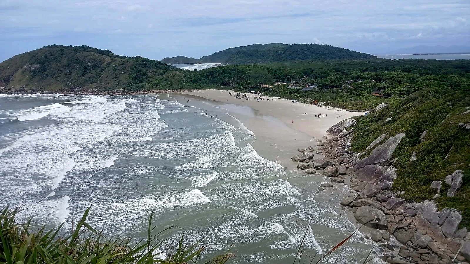 Vista elevada de uma praia de areia clara, com ondas quebrando junto a costões rochosos e mata densa ao redor; pequenas pessoas aparecem espalhadas pela faixa de areia. A cena tem luz diurna suave, sob céu parcialmente nublado, destacando o contraste