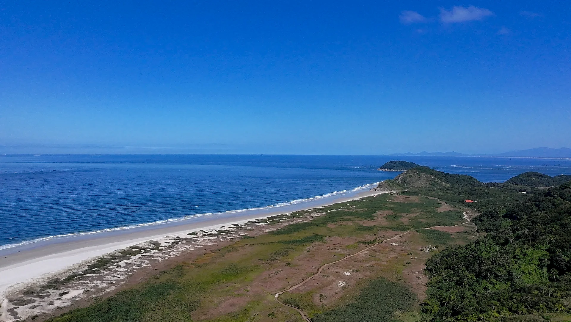 Vista aérea de uma praia de areia clara entre o mar azul e áreas de vegetação rasteira e mata densa, com morros ao fundo. A luz diurna intensa e o céu aberto destacam a paisagem costeira.