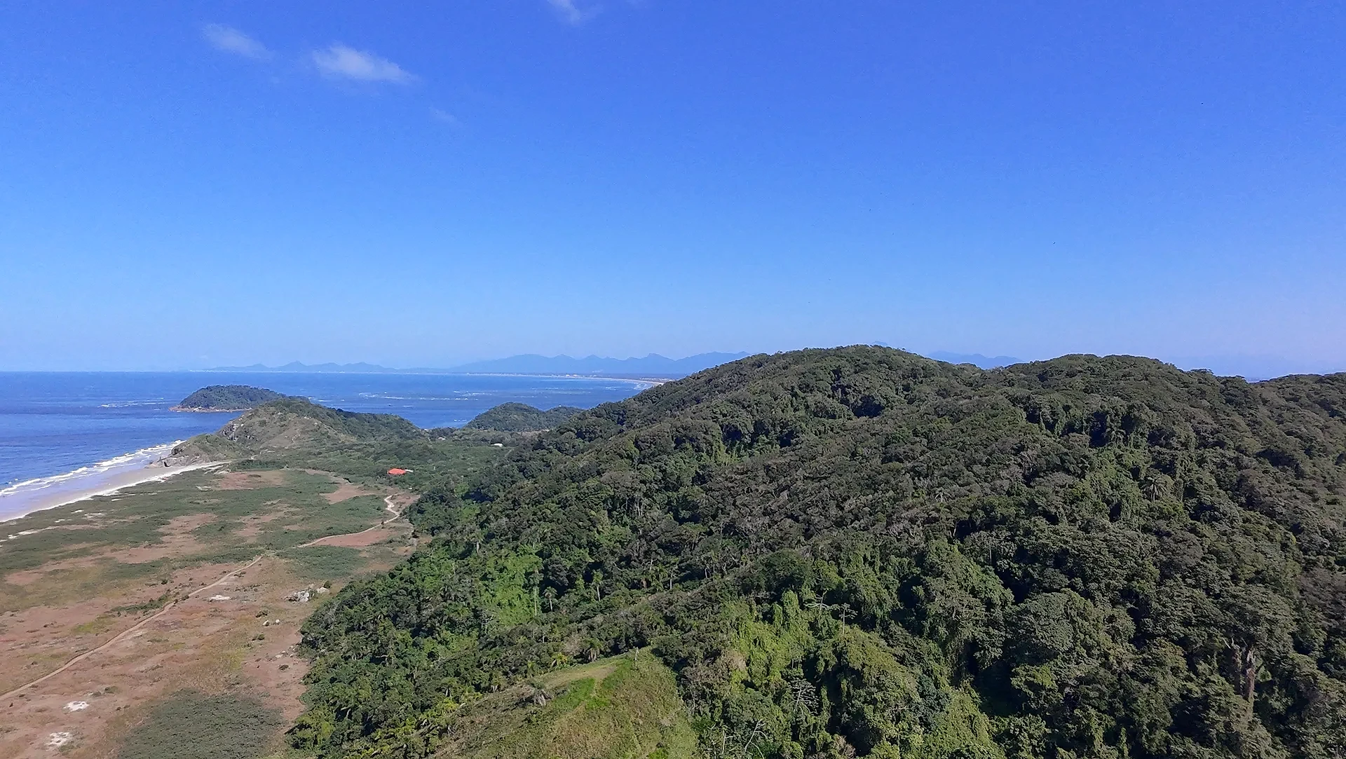 Vista aérea de uma praia extensa, com faixa de areia clara, mar azul e vegetação rasteira que avança até a mata. Uma trilha corta o terreno em direção à costa, sob luz solar intensa e céu limpo.