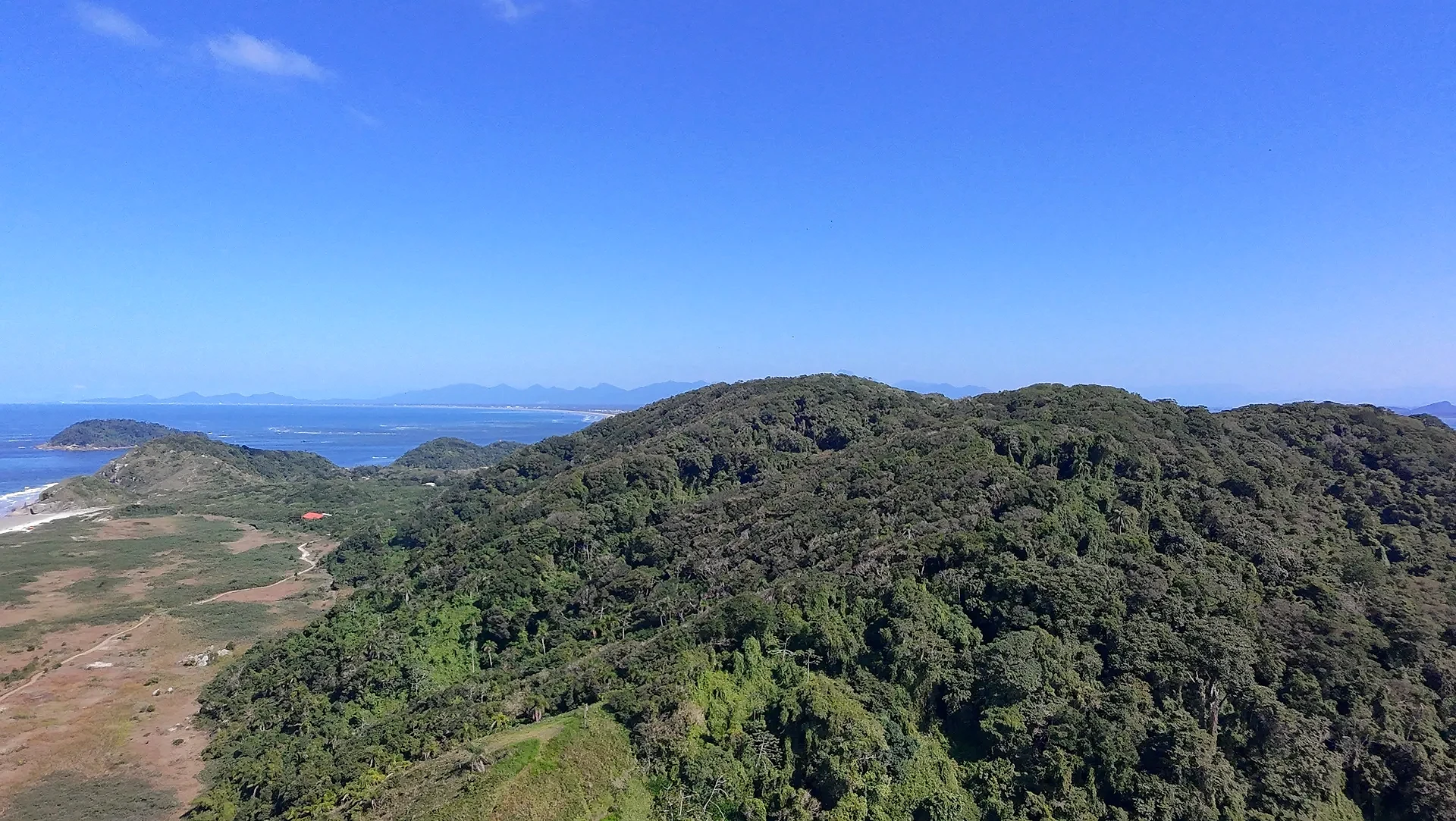 Vista aérea de uma praia extensa, com faixa de areia clara, mar azul e vegetação rasteira que avança até a mata. Uma trilha corta o terreno em direção à costa, sob luz solar intensa e céu limpo.