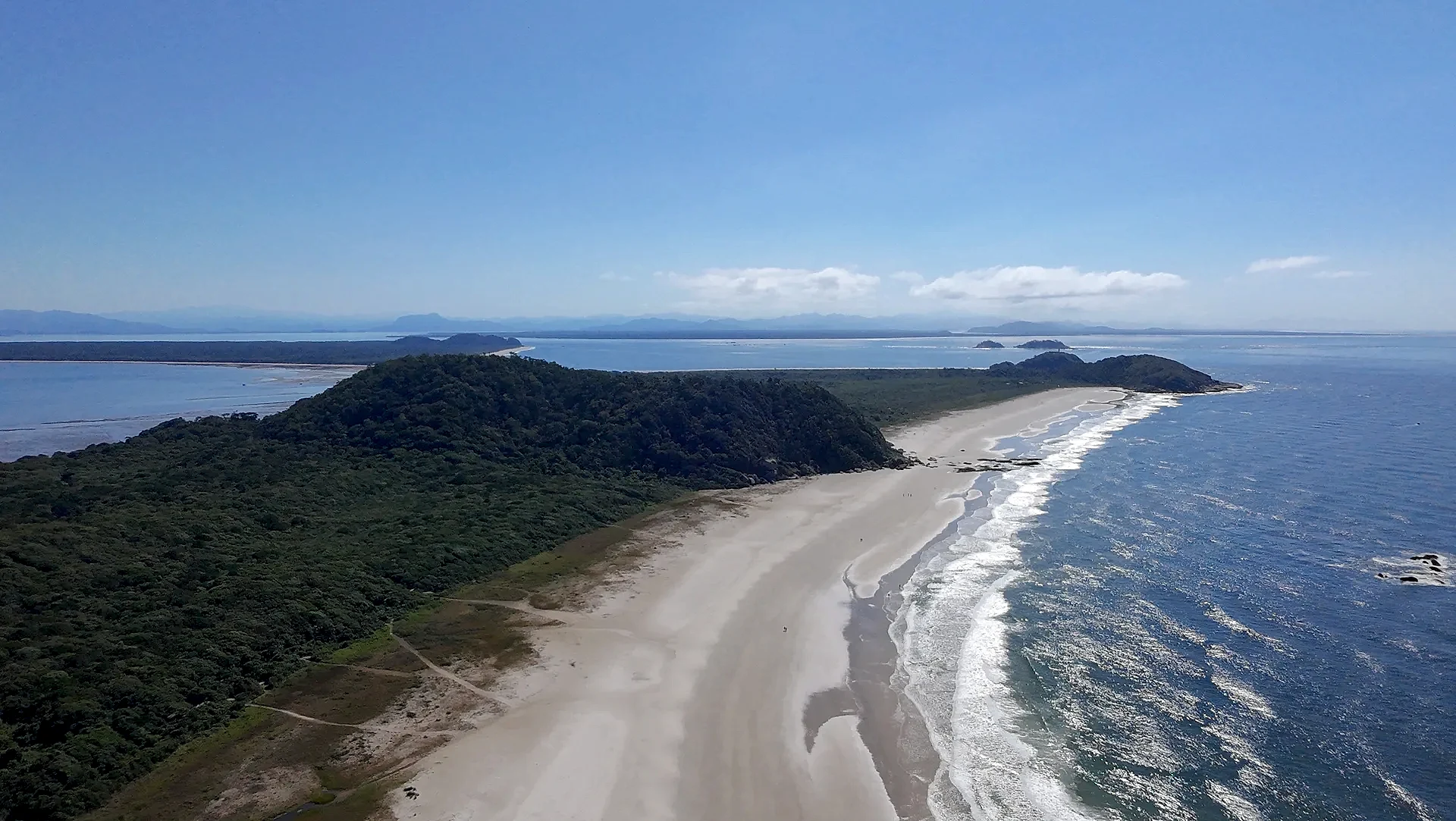 Vista aérea de uma praia extensa, com faixa de areia clara, mar azul e vegetação rasteira que avança até a mata. Uma trilha corta o terreno em direção à costa, sob luz solar intensa e céu limpo.