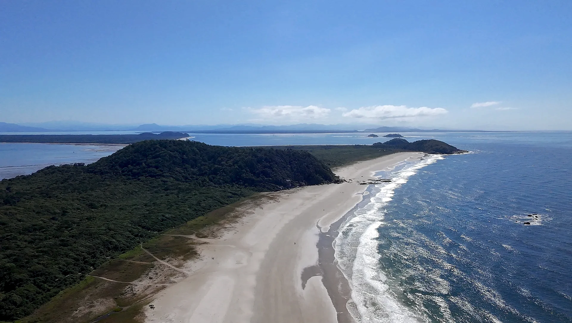 Vista aérea de uma praia extensa, com faixa de areia clara, mar azul e vegetação rasteira que avança até a mata. Uma trilha corta o terreno em direção à costa, sob luz solar intensa e céu limpo.