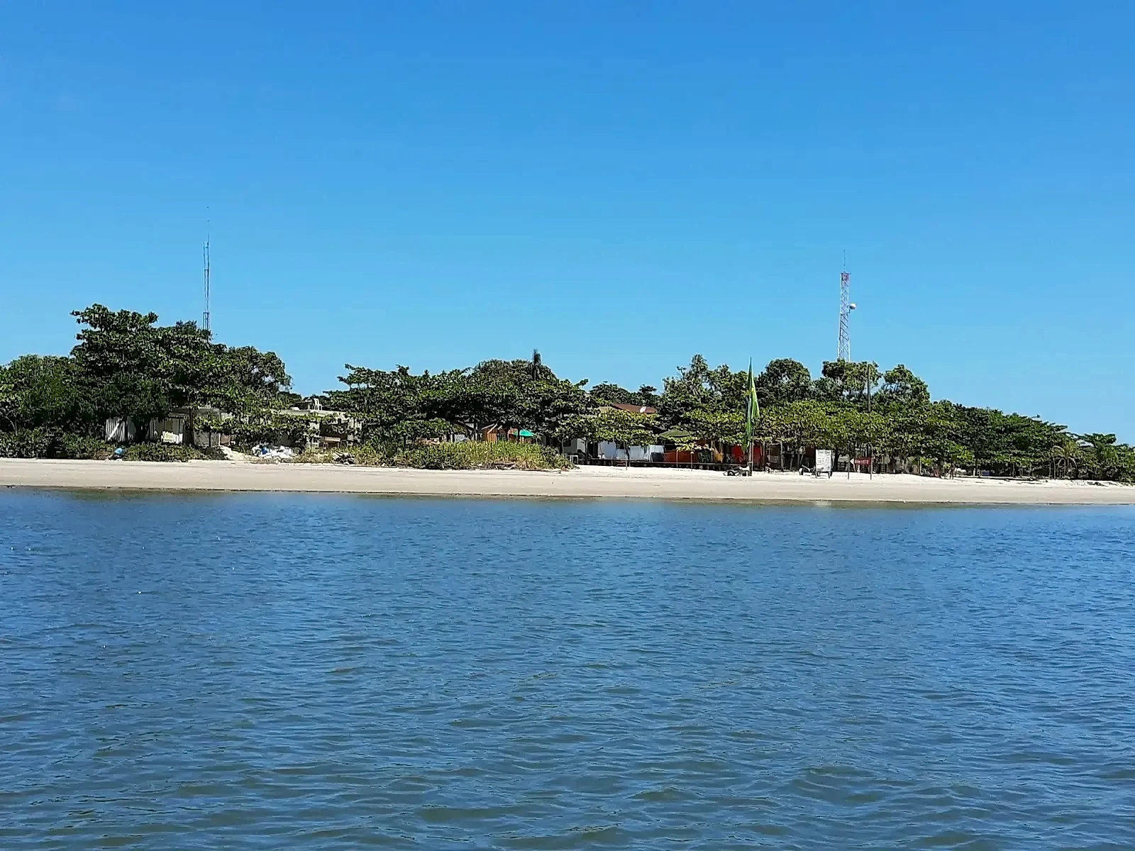 Faixa de praia de areia clara diante de águas calmas, com vila arborizada ao fundo. O céu azul aberto e a luz forte do dia destacam o ambiente litorâneo.