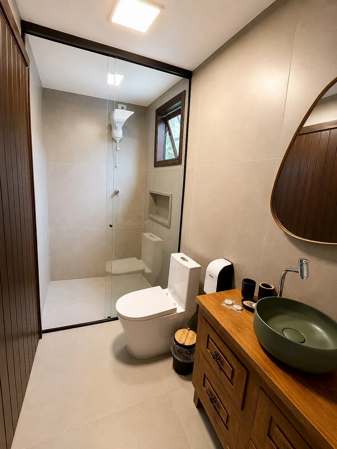 Banheiro compacto e bem iluminado, com box de vidro, revestimentos claros e bancada de madeira com cuba verde. A luz interna uniforme e a janela aberta criam um ambiente moderno e acolhedor.