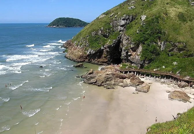 Praia de areia clara entre costões rochosos e mata verde, com pequenas ondas e algumas pessoas no mar. Um passarela de madeira acompanha as pedras sob luz solar intensa e céu aberto.