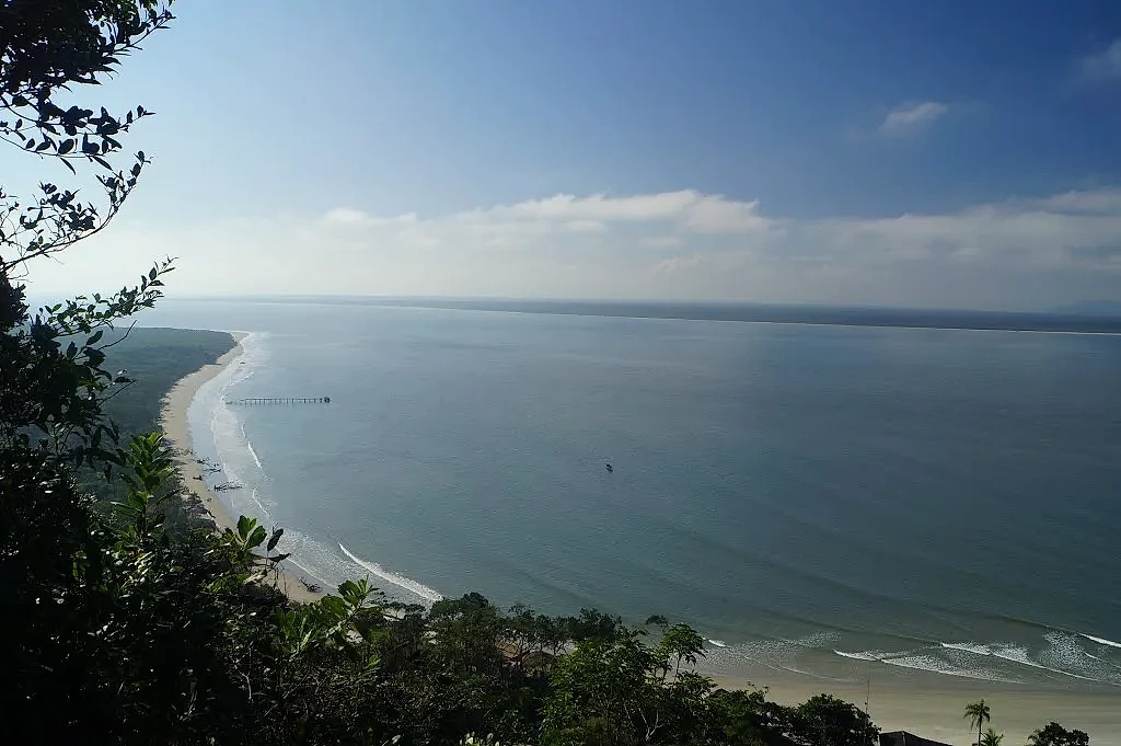 Vista elevada de uma praia de areia clara e mar calmo, contornada por mata e vegetação em primeiro plano. A luz diurna suave ilumina a costa sob céu azul com nuvens esparsas, e um pequeno trapiche aparece ao longe.