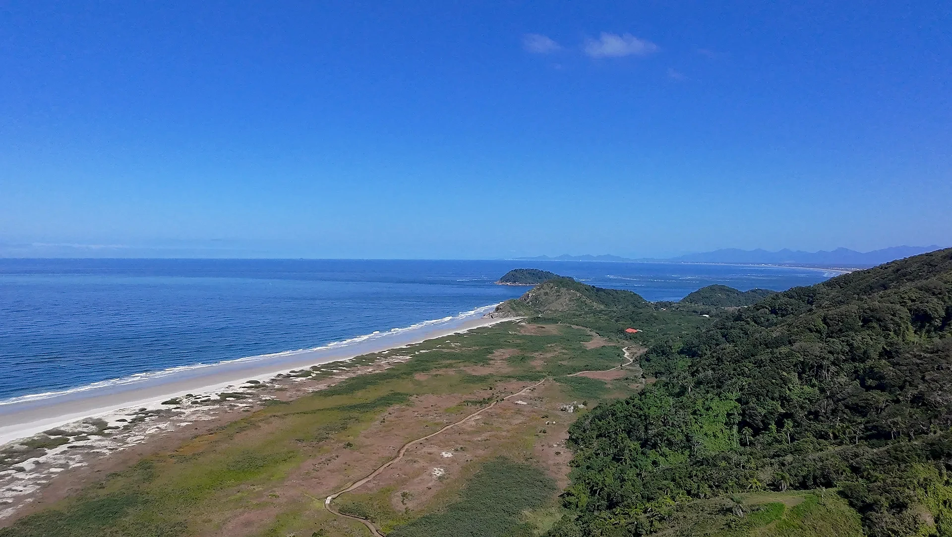 Vista aérea de uma praia extensa, com faixa de areia clara, mar azul e vegetação rasteira que avança até a mata. Uma trilha corta o terreno em direção à costa, sob luz solar intensa e céu limpo.