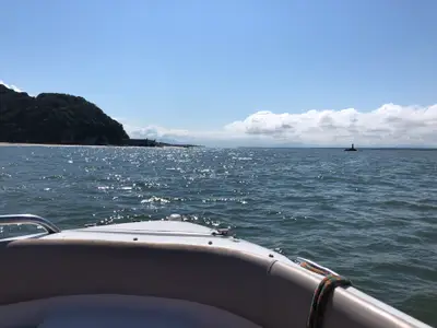 Vista a partir de um barco sobre o mar, com uma faixa de praia e mata densa ao fundo, além de um pequeno marco de navegação sobre as pedras. O dia é claro, com céu azul, nuvens no horizonte e luz solar intensa refletindo na água.