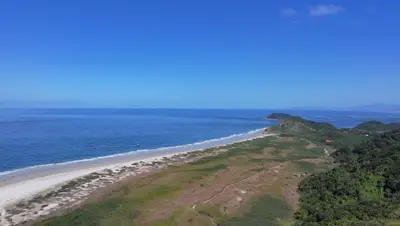 Vista aérea de uma praia de areia clara entre o mar azul e áreas de vegetação rasteira e mata densa, com morros ao fundo. A luz diurna intensa e o céu aberto destacam a paisagem costeira.