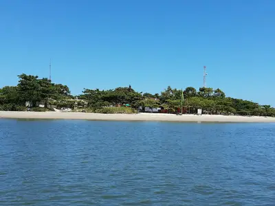 Faixa de praia de areia clara diante de águas calmas, com vila arborizada ao fundo. O céu azul aberto e a luz forte do dia destacam o ambiente litorâneo.