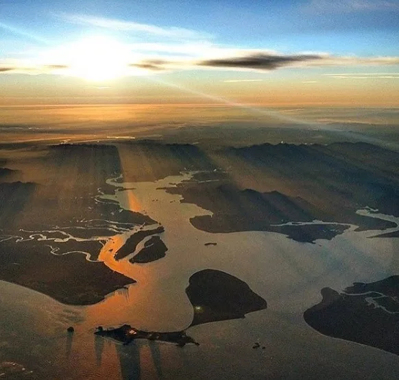 Vista aérea de canais, ilhas e áreas de mata costeira banhados pela luz dourada do entardecer. O céu parcialmente nublado e os reflexos alaranjados na água criam uma atmosfera serena.