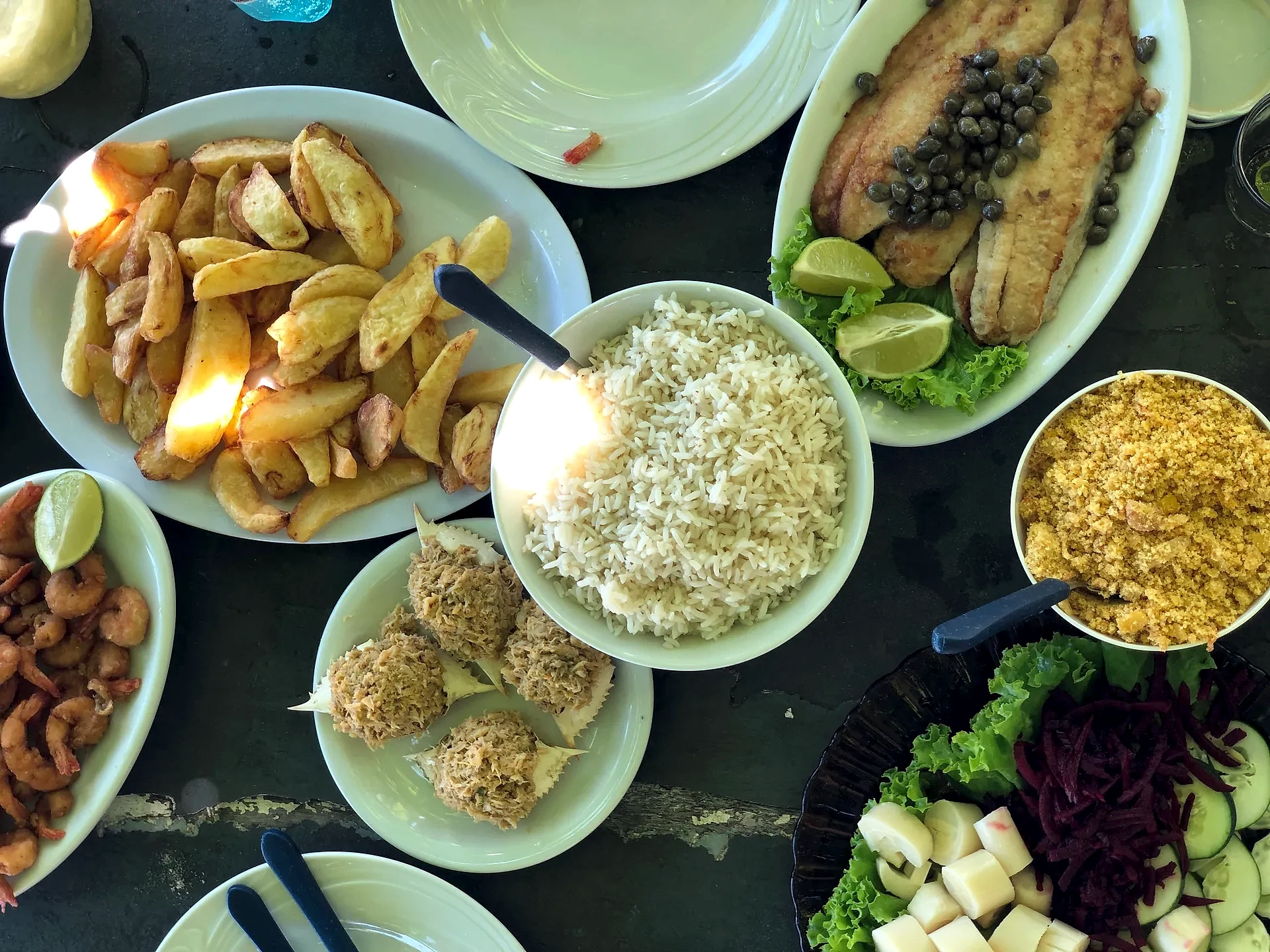 Mesa com pratos de peixe frito, camarão, arroz, farofa, batatas e salada, compondo uma refeição típica à base de frutos do mar. A cena é vista de cima, em ambiente descontraído, com luz natural incidindo sobre os alimentos.