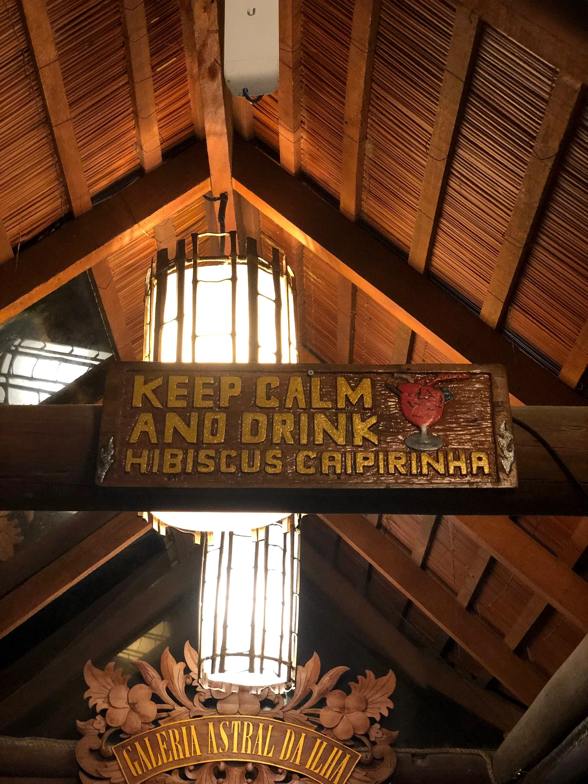 Interior rústico com teto de bambu, luminárias e placas decorativas de madeira em um ambiente de hospedagem ou restaurante. A iluminação quente cria uma atmosfera acolhedora e intimista.