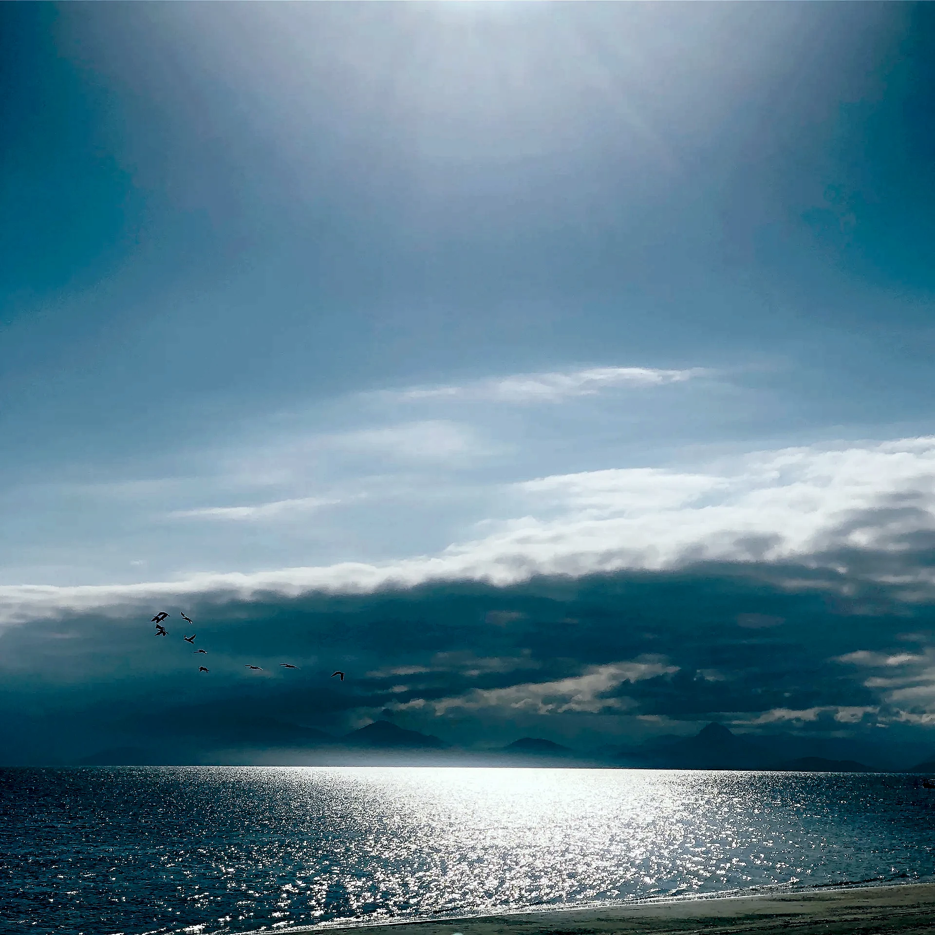 Praia de mar calmo sob céu amplo e parcialmente nublado, com aves sobrevoando ao longe e montanhas suaves no horizonte. A luz clara do dia se reflete intensamente na água, criando um brilho prateado sobre o mar.
