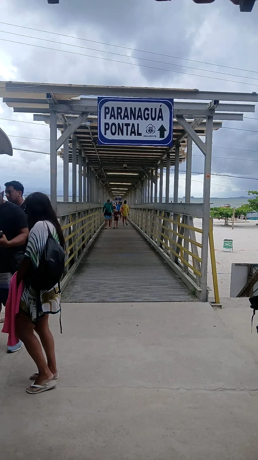 Trapiche coberto com pessoas caminhando em direção ao embarque, com praia e baía ao fundo. O céu nublado cria uma luz suave e difusa sobre o ambiente costeiro.
