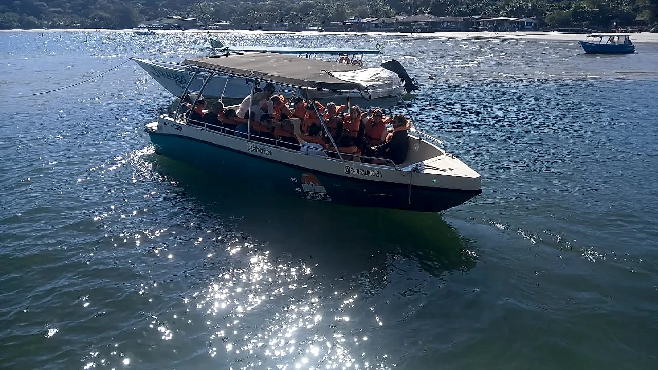 Barco de passageiros navega em águas calmas diante de uma praia com mata e construções à beira-mar. A luz forte do dia reflete na superfície da baía, criando brilhos sobre a água.