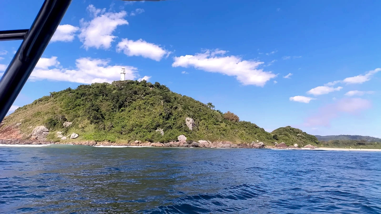 Farol no alto de um morro coberto por mata, visto do mar a partir de um barco, com costões rochosos e pequena faixa de praia ao fundo. A cena tem luz solar intensa, céu azul e nuvens esparsas.