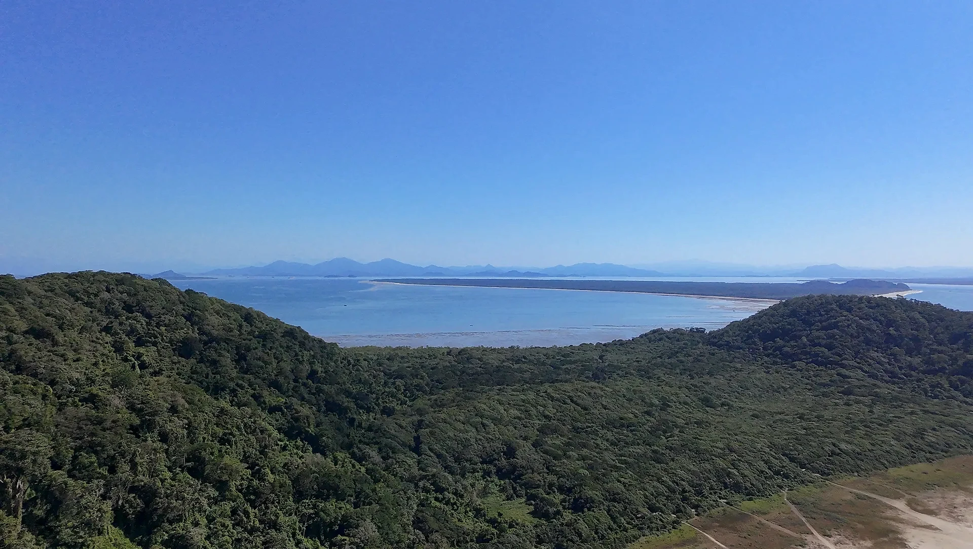 Vista aérea de uma praia extensa, com faixa de areia clara, mar azul e vegetação rasteira que avança até a mata. Uma trilha corta o terreno em direção à costa, sob luz solar intensa e céu limpo.