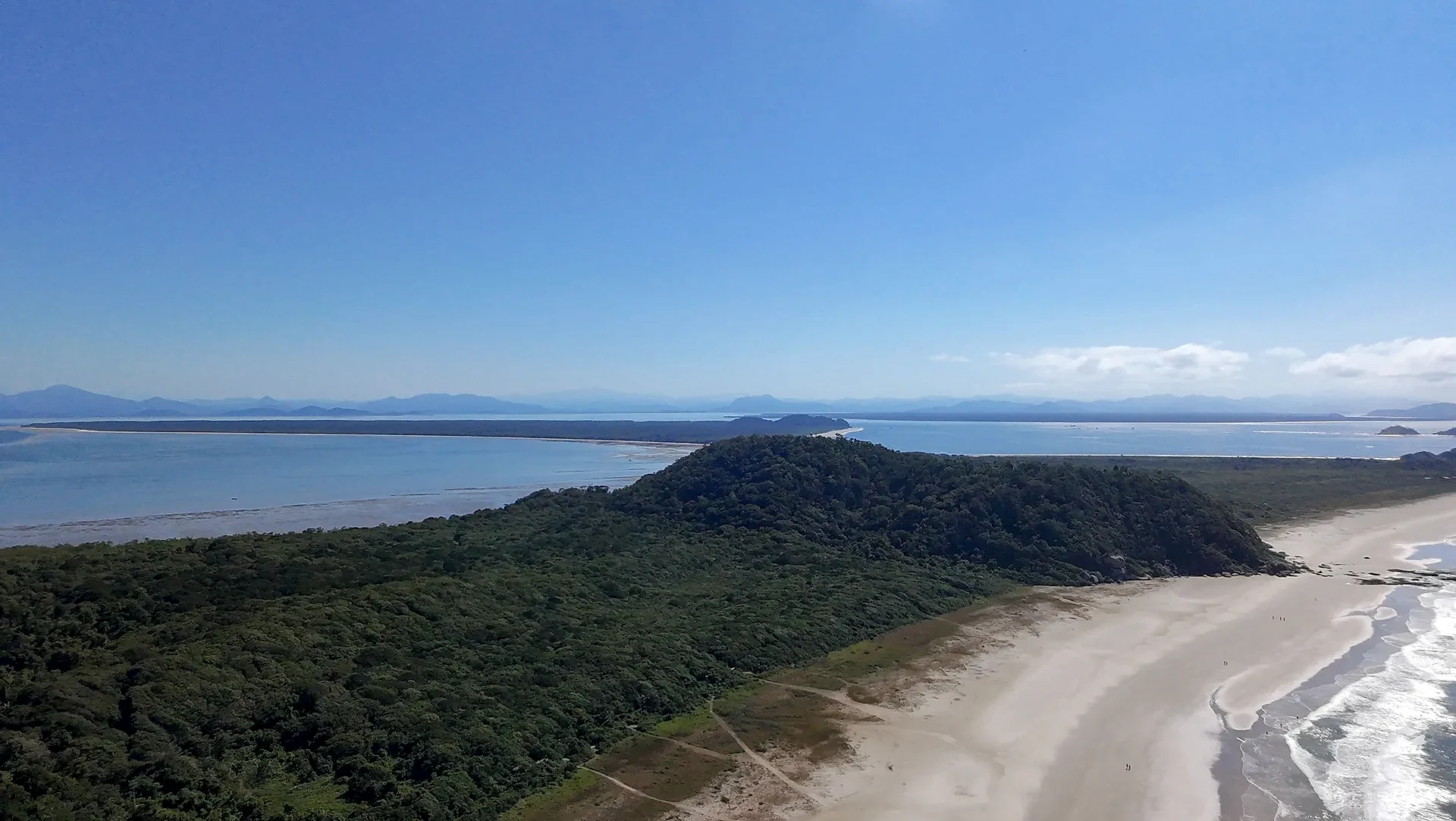 Vista aérea de uma praia extensa, com faixa de areia clara, mar azul e vegetação rasteira que avança até a mata. Uma trilha corta o terreno em direção à costa, sob luz solar intensa e céu limpo.