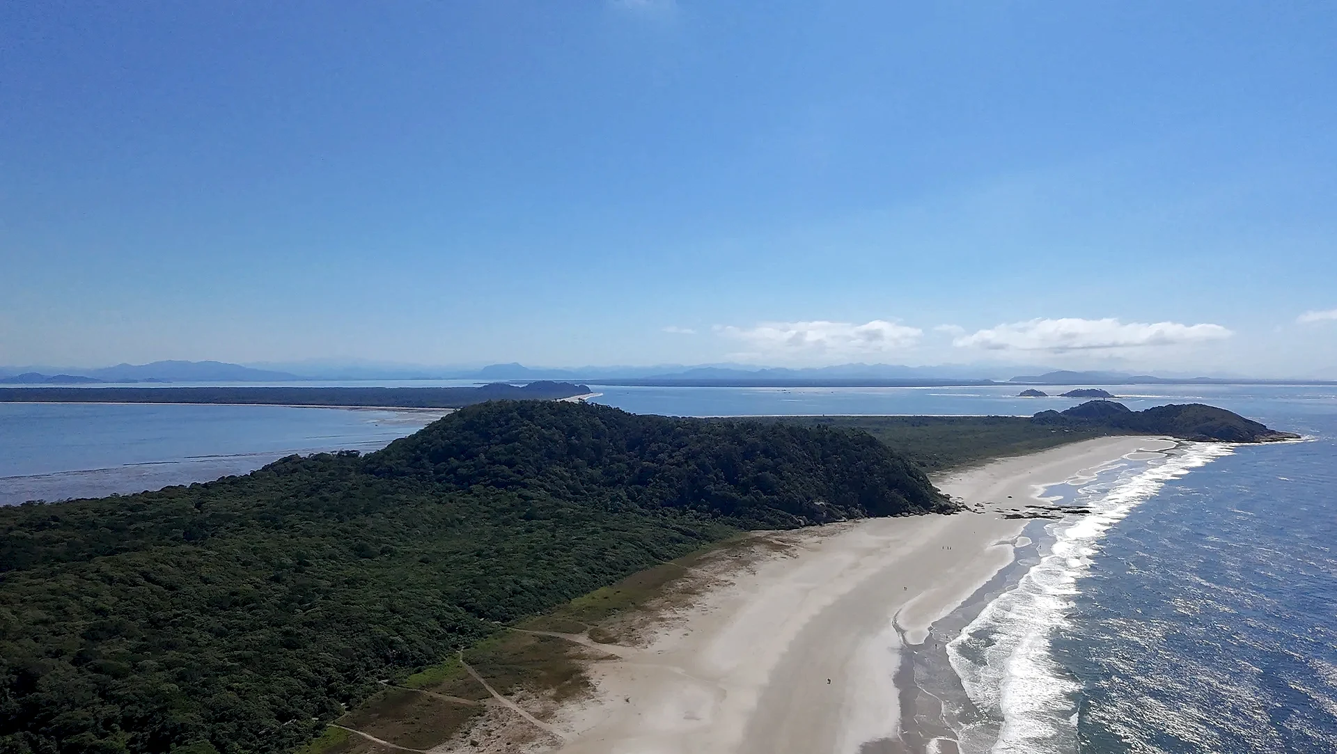 Vista aérea de uma praia extensa, com faixa de areia clara, mar azul e vegetação rasteira que avança até a mata. Uma trilha corta o terreno em direção à costa, sob luz solar intensa e céu limpo.