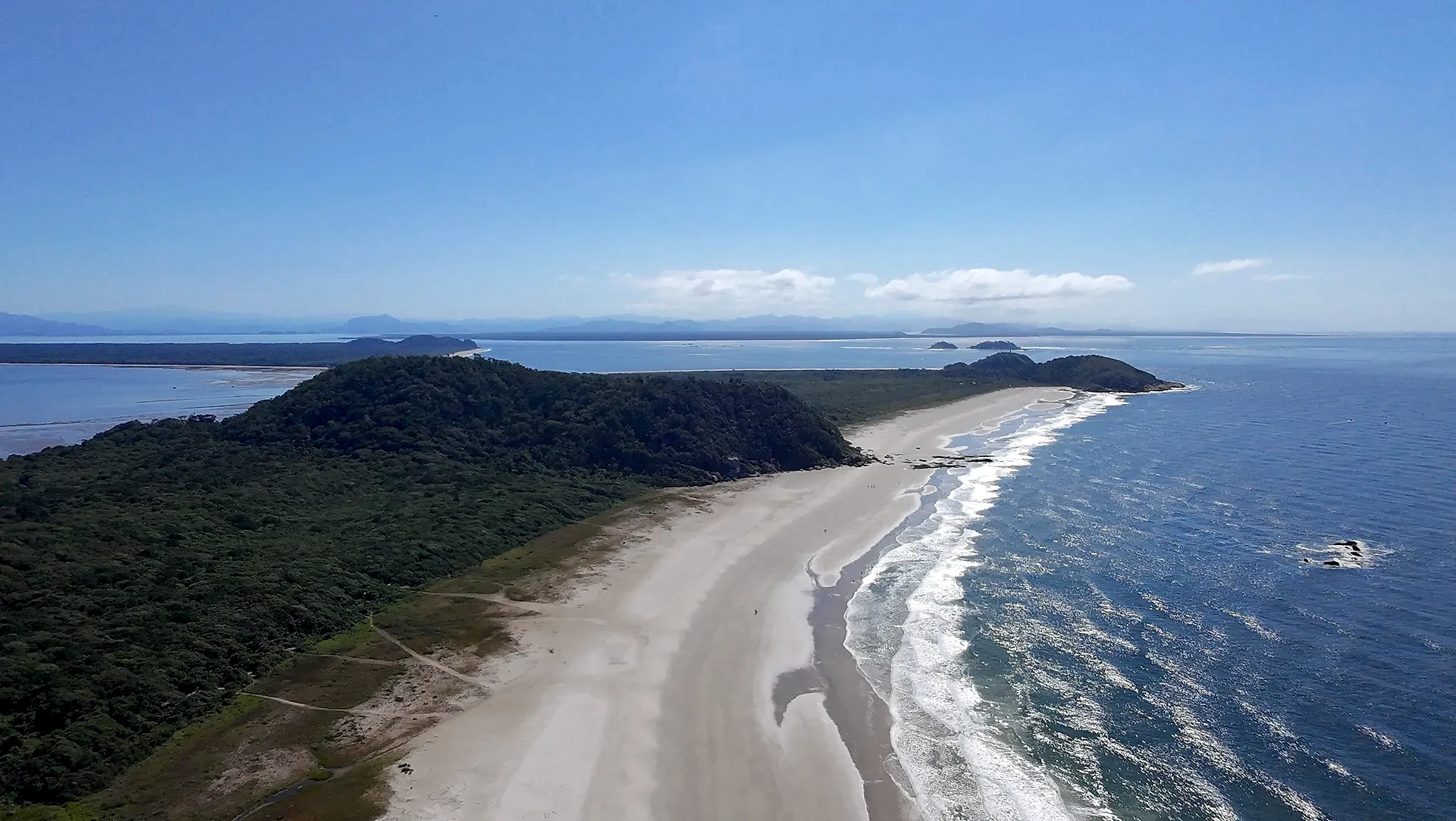 Vista aérea de uma praia extensa, com faixa de areia clara, mar azul e vegetação rasteira que avança até a mata. Uma trilha corta o terreno em direção à costa, sob luz solar intensa e céu limpo.
