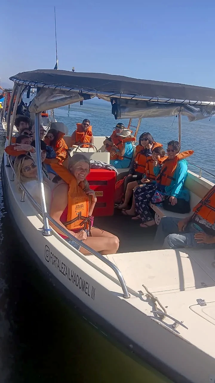Grupo de pessoas com coletes salva-vidas em barco de passeio sobre águas calmas, sob cobertura de lona. A cena é iluminada por sol forte, com céu azul e clima de passeio marítimo.